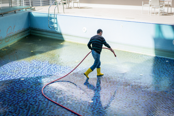 A man in yellow rubber boots cleaning a pool with a brush on a sunny day.